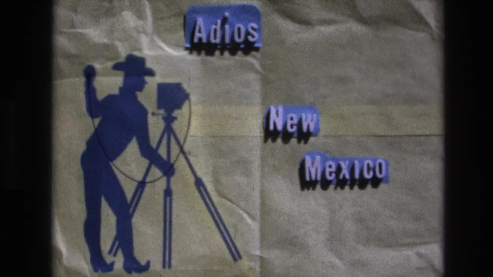 Final extracted 1973 New Mexico frame showing the handmade brown-paper farewell card with a blue cowboy photographer silhouette, tripod camera, and cut-out "Adios New Mexico" text.