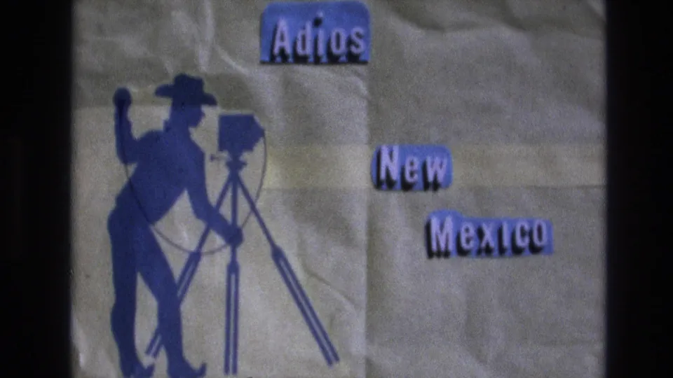 1973 New Mexico home-movie title card frame with blue Adios New Mexico lettering, a cowboy photographer silhouette, tripod camera, paper texture, and dark 8mm border.