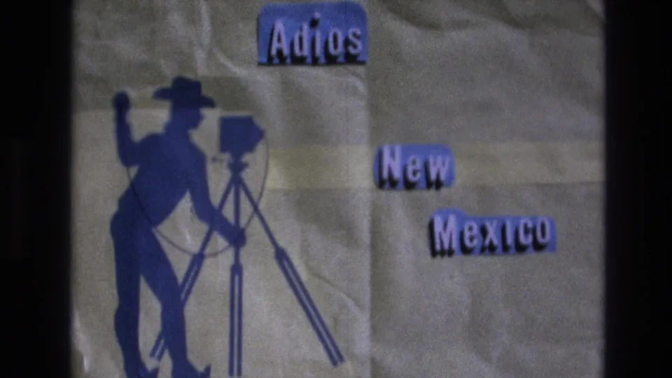 1973 New Mexico travel title card with cut-out Adios New Mexico lettering and a blue cowboy figure standing beside a tripod-mounted camera.