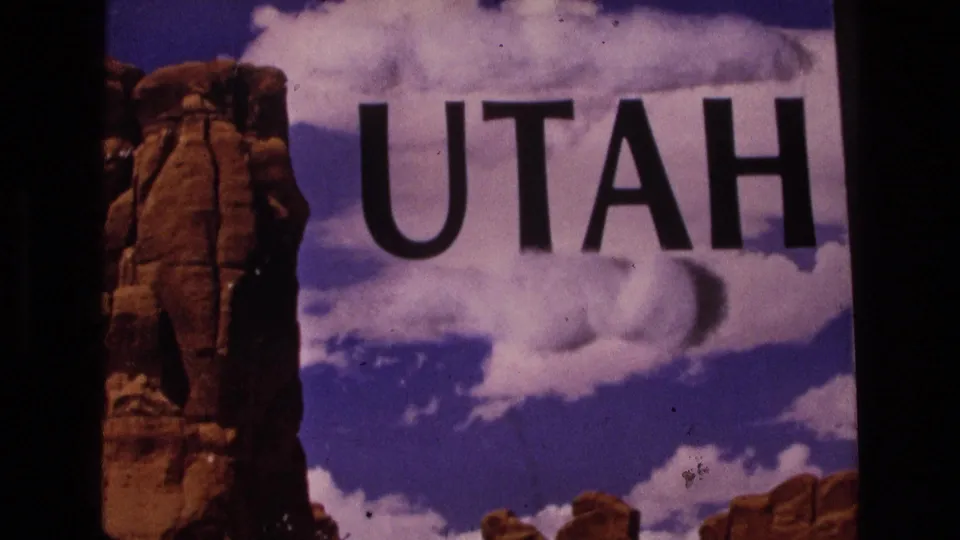 1973 8mm frame of a Utah place-title graphic, with large black letters set against clouds, blue sky, and reddish rock scenery.