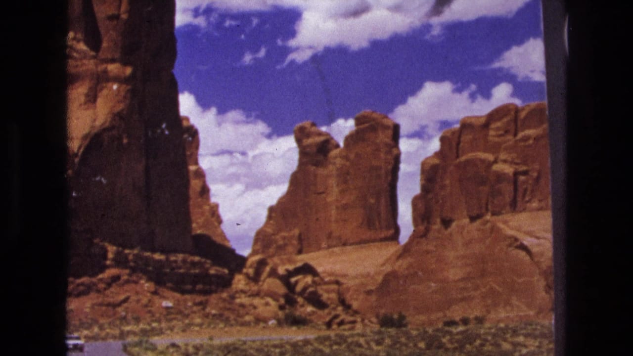 Red rock desert formations beside a paved road under a blue, clouded sky.