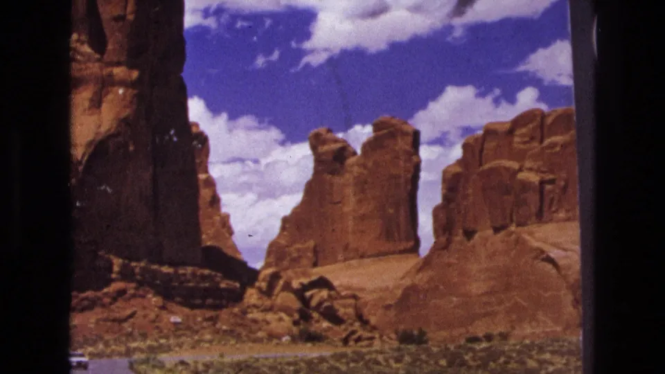 1970s 8mm frame of a red rock desert landscape with sandstone towers, scrub terrain, and a paved road seen from a moving travel perspective.