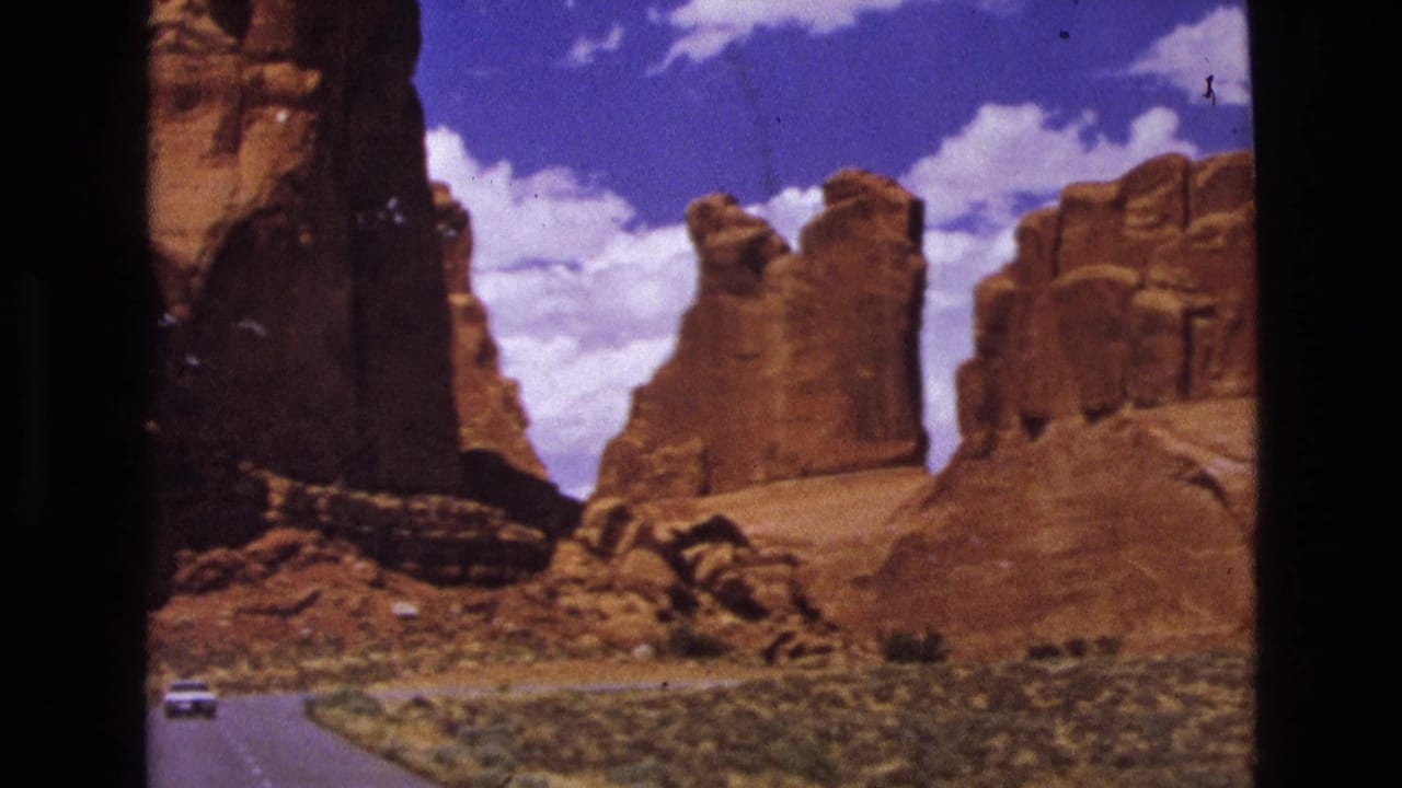 1970s 8mm view of a paved road curving through red sandstone terrain, with a small white car and cloud-filled blue sky visible.