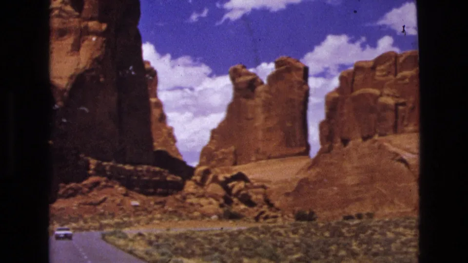 1970s 8mm view of a paved road curving through red sandstone terrain, with a small white car and cloud-filled blue sky visible.