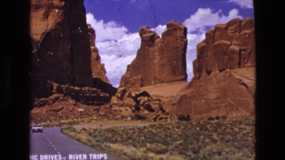 1970s 8mm frame showing a white car on a desert highway below red sandstone towers, with partial text reading scenic drives and river trips at the bottom edge.