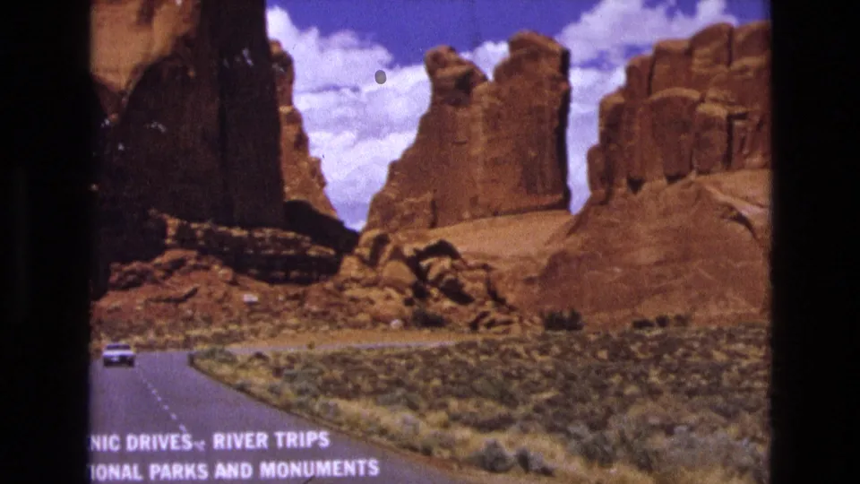 1970s 8mm travel frame of a winding desert road, sandstone formations, a distant white car, and bottom-edge text referencing scenic drives, river trips, national parks, and monuments.
