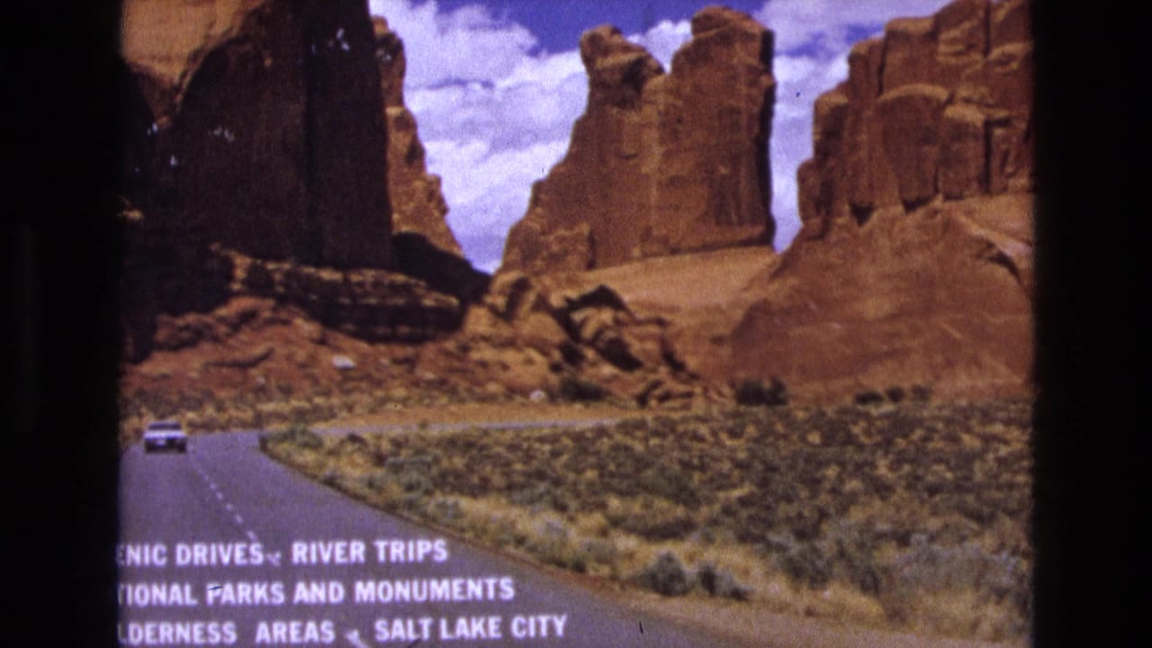Paved desert road leading toward red sandstone cliffs with a small car in the distance and white travel text at the bottom.