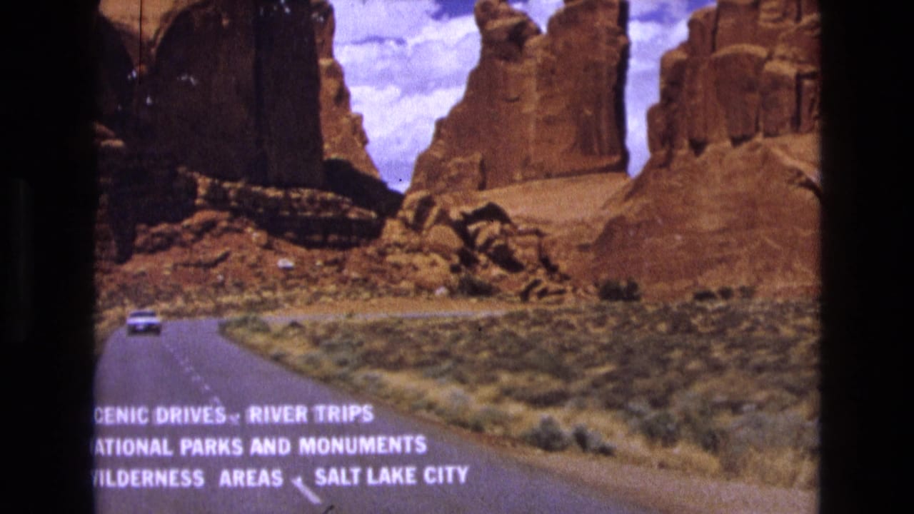 1973 8mm frame of a highway approaching red sandstone formations in Utah, with a small car ahead and lower-frame text promoting scenic drives, national parks, wilderness areas, and Salt Lake City.