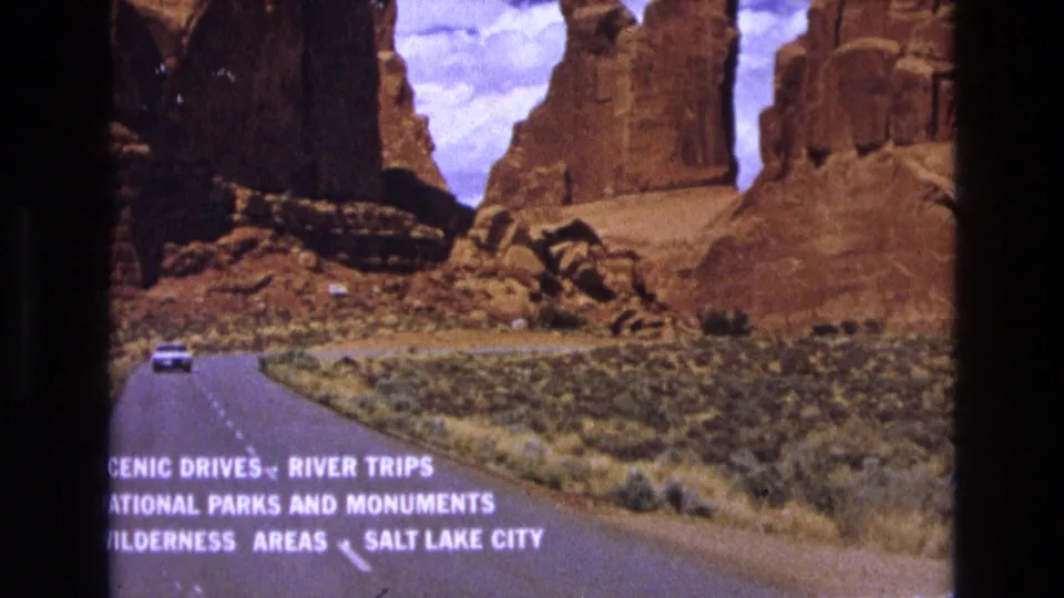 Vintage 8mm frame from the 1973 Utah travel clip showing a curving road through sparse desert vegetation toward red rock cliffs, with promotional travel wording visible across the bottom.