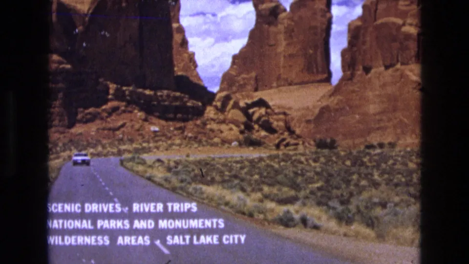 1973 8mm Utah road-trip frame showing a distant vehicle on a paved desert road beneath red sandstone formations, with white text referencing scenic drives and Salt Lake City.