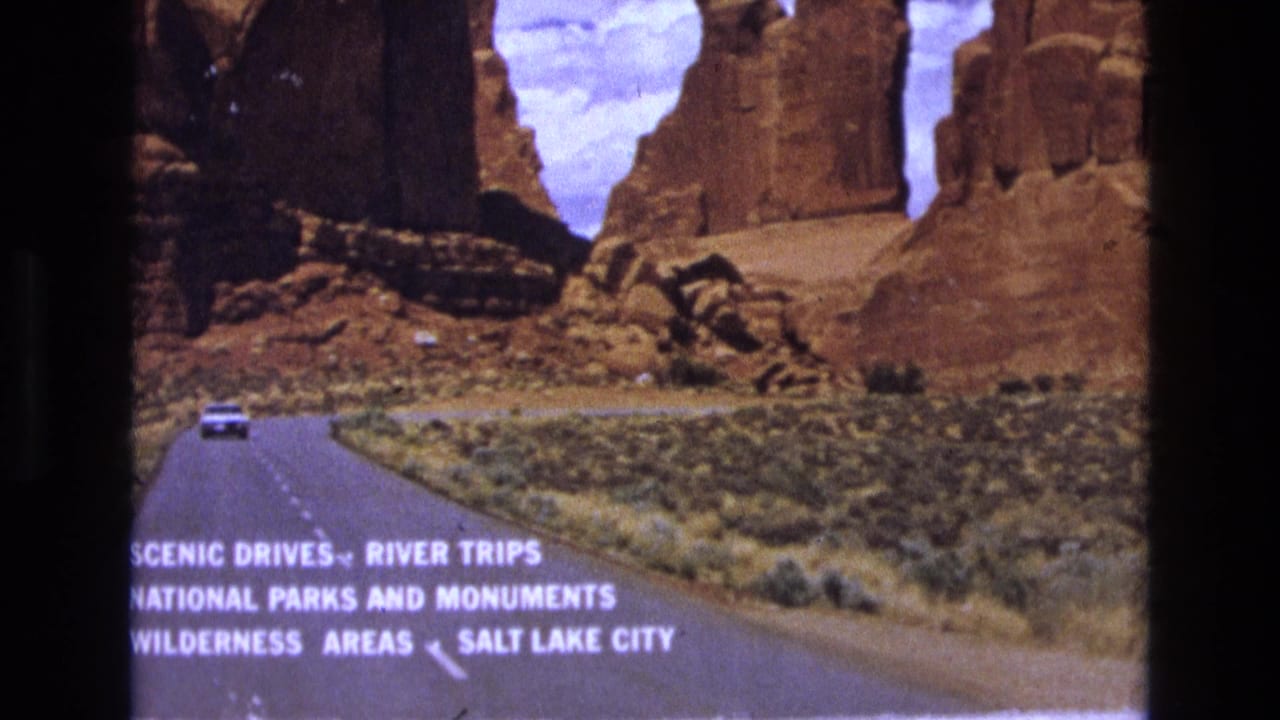1973 8mm home-movie frame showing a white car on a Utah canyon road, red sandstone formations, desert brush, and directional travel text pointing toward Salt Lake City.