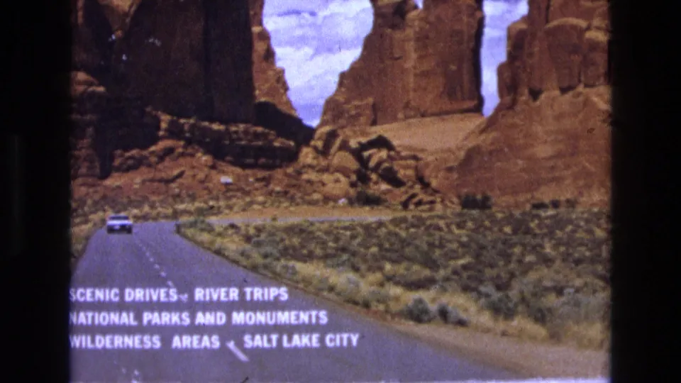 1973 8mm home-movie frame showing a white car on a Utah canyon road, red sandstone formations, desert brush, and directional travel text pointing toward Salt Lake City.