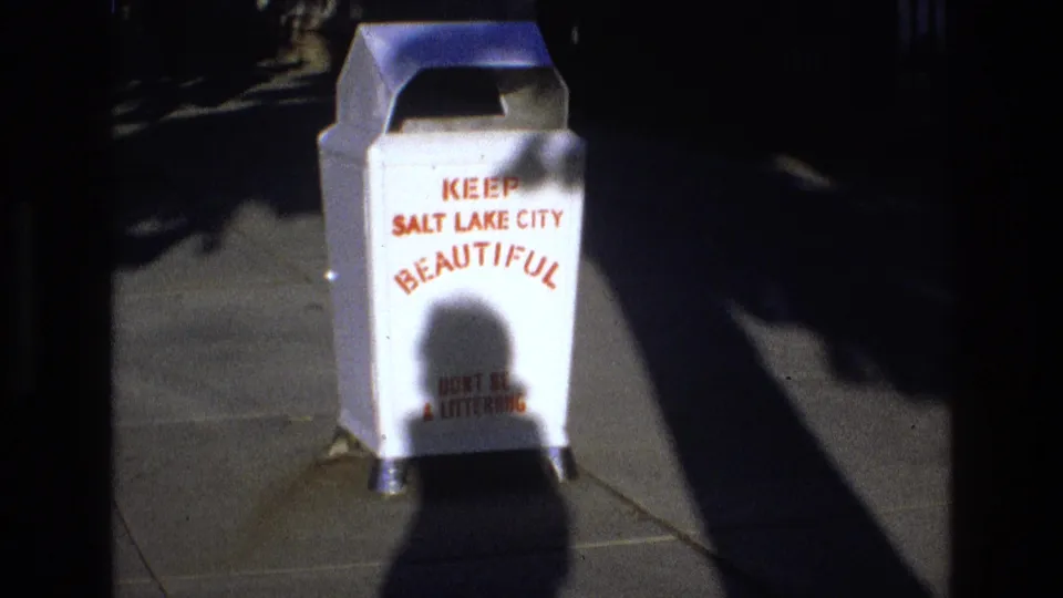 1973 Salt Lake City street cutaway showing a white public trash can on a sidewalk with red Keep Salt Lake City Beautiful lettering and deep sun shadows.