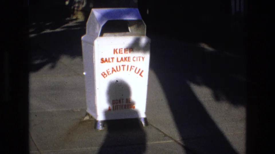 1973 Salt Lake City home-movie frame holding on a white civic trash can with Keep Salt Lake City Beautiful text, sidewalk paving, and high-contrast afternoon shadows.