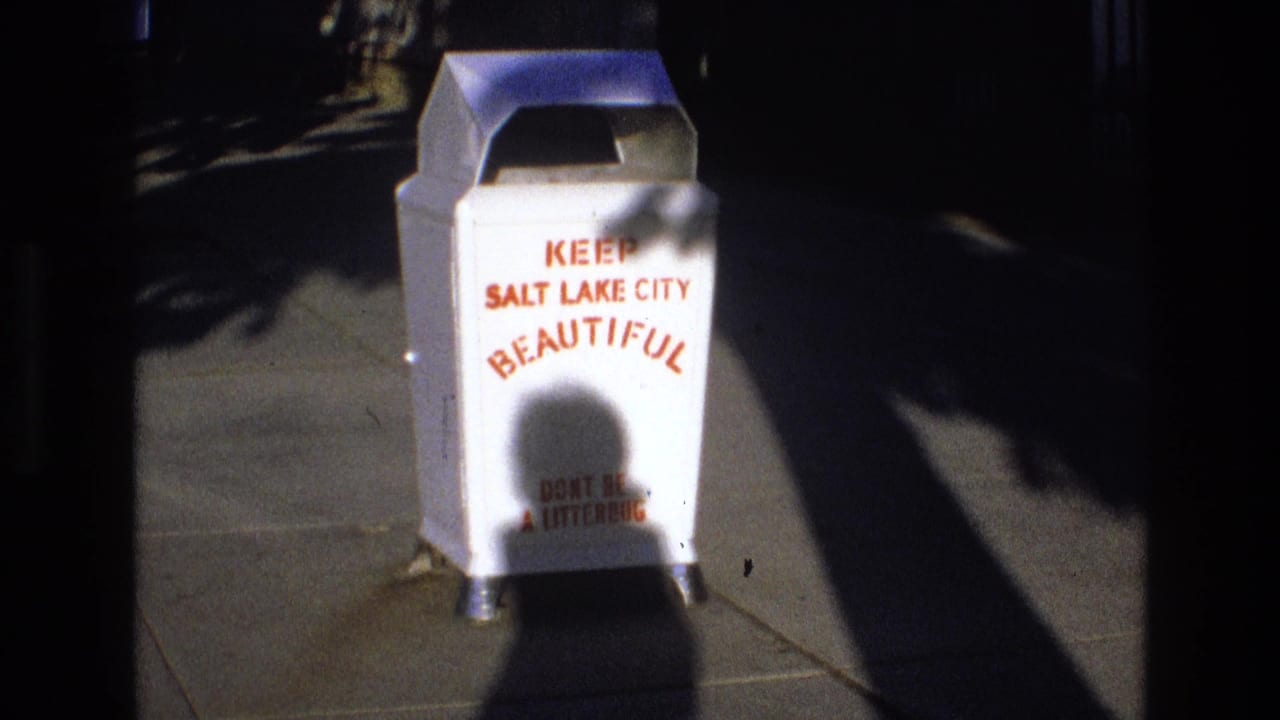 1973 Salt Lake City home-movie frame showing a white public litter container reading "Keep Salt Lake City Beautiful" on a sidewalk in strong shadow.