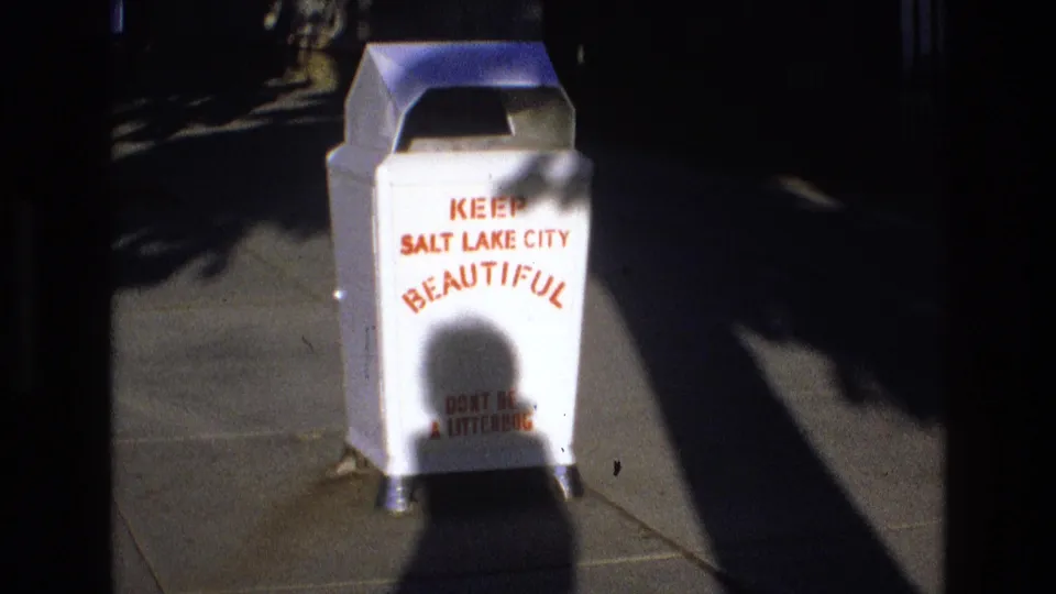 1973 Salt Lake City home-movie frame showing a white public litter container reading "Keep Salt Lake City Beautiful" on a sidewalk in strong shadow.