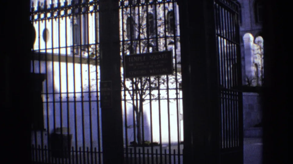 1973 Salt Lake City home-movie frame showing the Temple Square sign behind ornate black fencing, with a tree and light-toned buildings in the background.