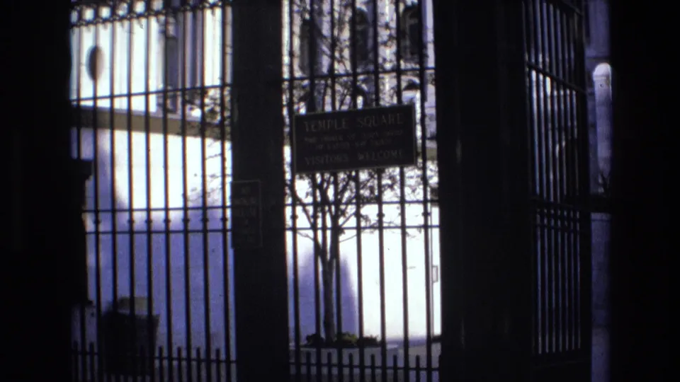 1973 Salt Lake City home-movie frame looking through a wrought iron gate toward a Temple Square visitors welcome sign and smaller no smoking notice.