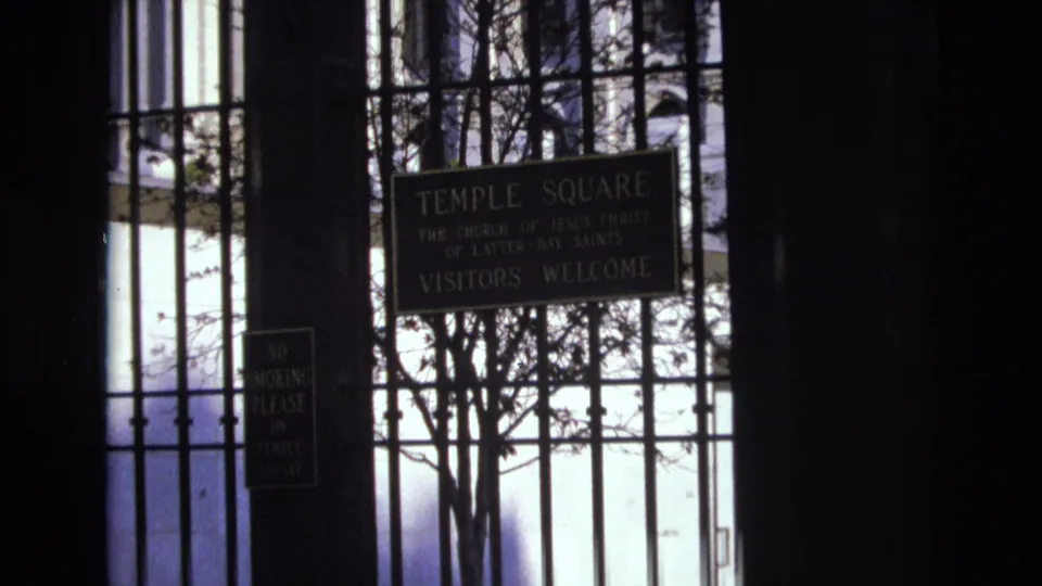 1973 Salt Lake City frame with the camera zoomed closer on the Temple Square sign reading visitors welcome, partially framed by gate bars and tree branches.