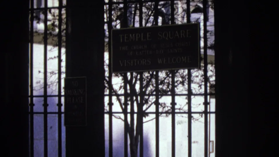 Final 1973 Salt Lake City frame holding on the Temple Square visitors welcome sign behind wrought iron bars, with tree branches crossing the bright background.
