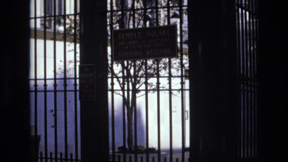 1973 Salt Lake City frame looking through the Temple Square gate toward a sign welcoming visitors, with bare trees silhouetted against a bright background.
