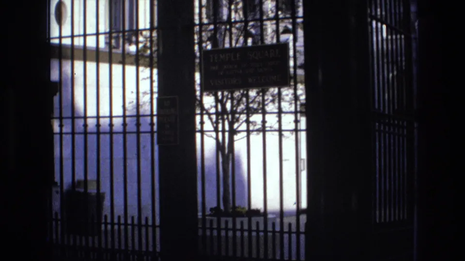 1973 Salt Lake City home-movie frame showing the Temple Square gate from a visitor perspective, with signage, vertical bars, and leafless trees in high-contrast light.