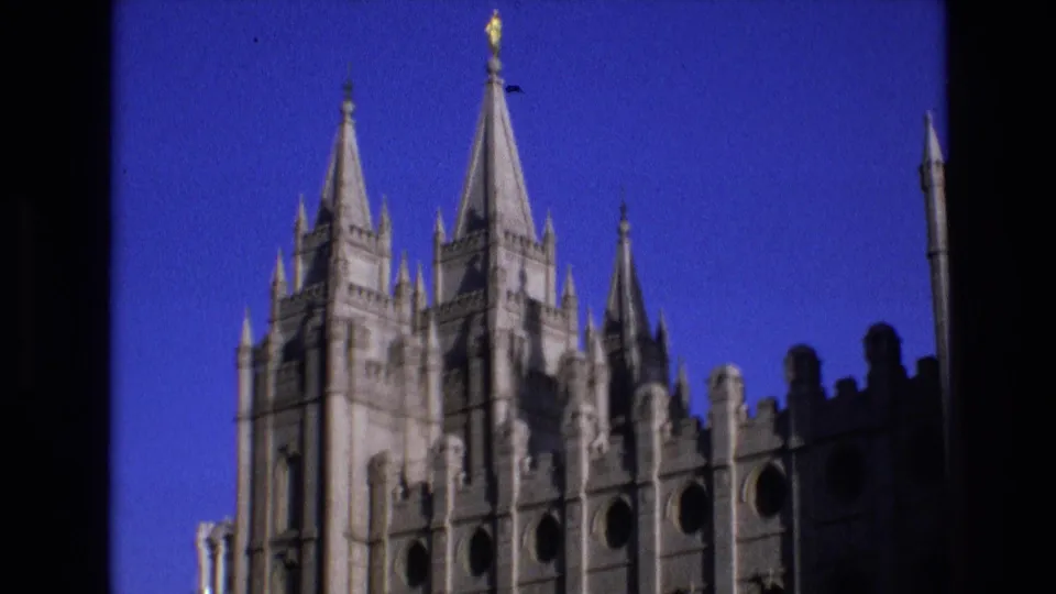 Visitor-shot 1973 view of the Salt Lake City Temple upper facade, showing tall stone spires, crenellations, circular openings, and strong blue sky color.