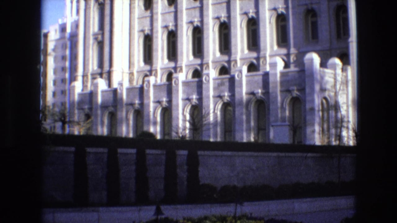 Salt Lake City temple exterior in 1973, with repeated arched windows, pale stonework, a perimeter wall, shrubs, and dark framing at the edge of the home-movie shot.