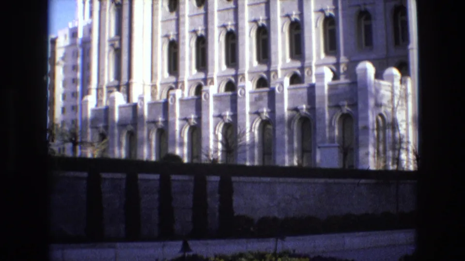 Salt Lake City temple exterior in 1973, with repeated arched windows, pale stonework, a perimeter wall, shrubs, and dark framing at the edge of the home-movie shot.