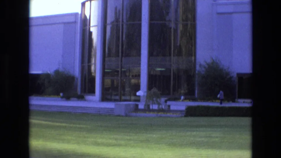 1973 8mm Salt Lake City view of a modern building entrance with tall glass panels, white columns, steps, landscaping, and a broad green lawn.