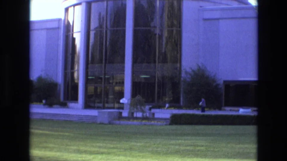 Salt Lake City 1973 home-movie frame showing a modern glass-fronted building, landscaped lawn, steps, columns, shrubs, and an incidental pedestrian at a distance.