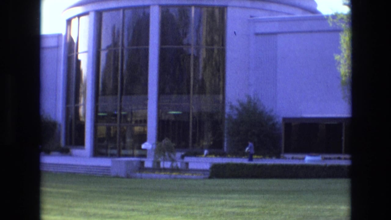 Distant person outside a curved glass-front building with white columns and lawn.