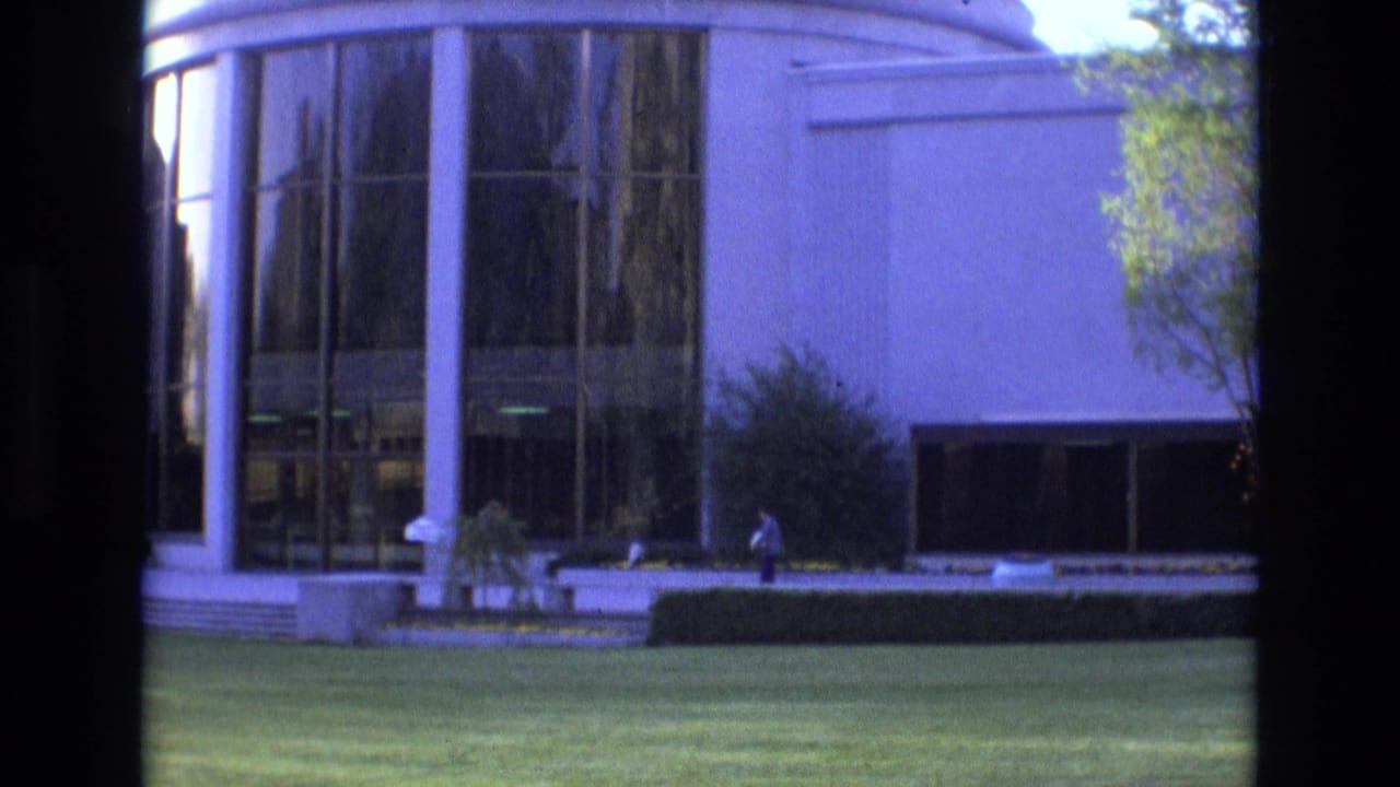 1973 Salt Lake City 8mm frame with a distant waiting figure in front of a modern curved glass facade, white exterior walls, landscaping, and strong dark borders from the film or viewing frame.