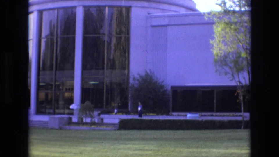 1973 Salt Lake City frame showing a white-columned modern building, reflective glass, clipped lawn, shrubs, and one distant person standing near the walkway or entrance area.