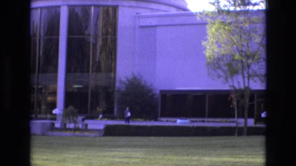 1973 Salt Lake City 8mm frame with a distant figure near a landscaped modern building exterior, including glass panels, white walls, hedges, trees, and a broad green lawn.