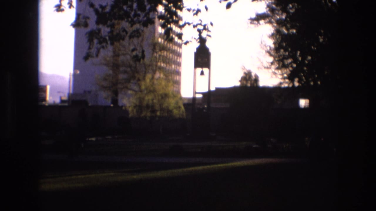 1973 Salt Lake City home-movie frame with a silhouetted bell tower, tall windowed building, tree branches, and dark foreground framing against a bright sky.