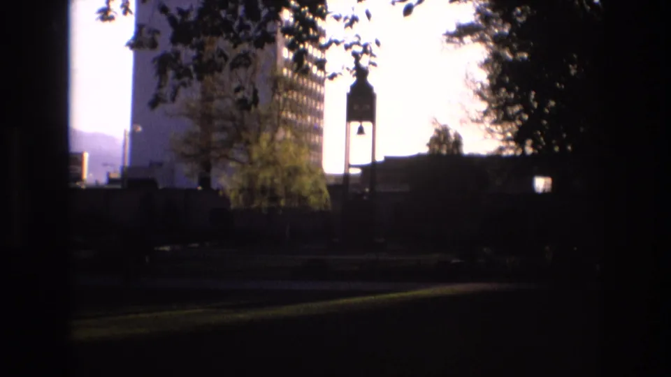 1973 Salt Lake City home-movie frame with a silhouetted bell tower, tall windowed building, tree branches, and dark foreground framing against a bright sky.