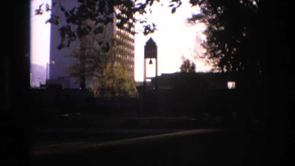 Backlit 1973 Salt Lake City exterior view showing a bell-tower-like structure beside a tall building, with tree canopy and distant hills partially visible.