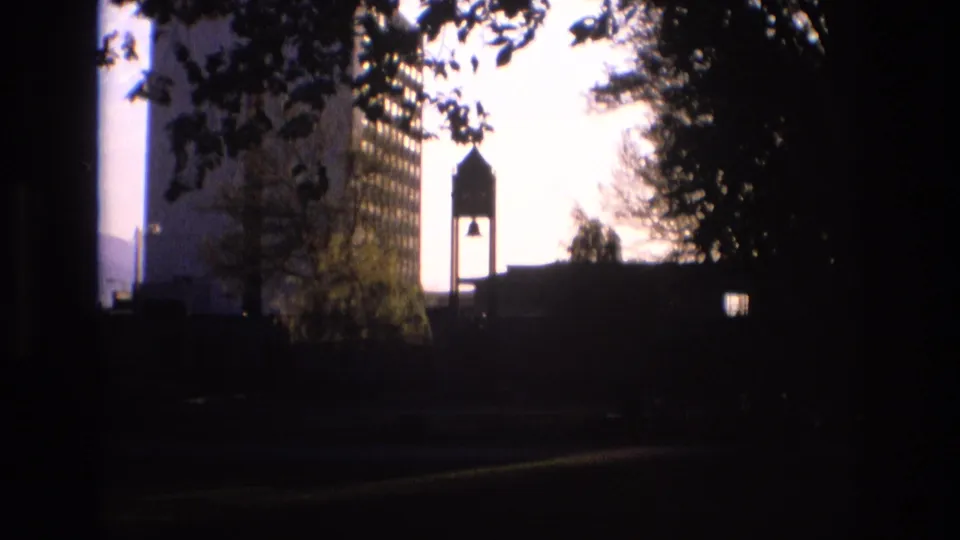 Soft 8mm frame from Salt Lake City in 1973 showing a tall modern building, silhouetted bell tower, trees, and a dark walkway or lawn in the foreground.