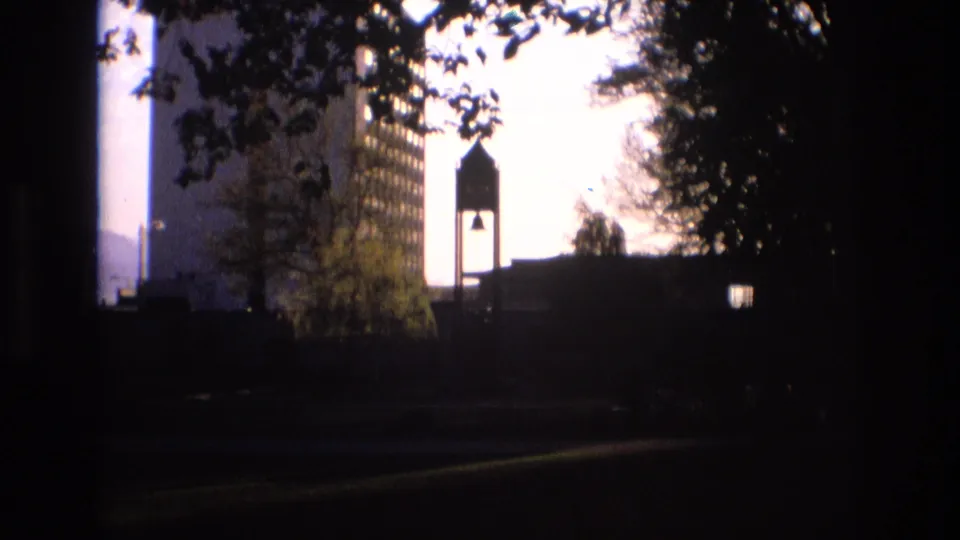 1973 Salt Lake City home-movie still with the bell tower and tall building remaining in silhouette, framed by tree branches and heavy shadow.