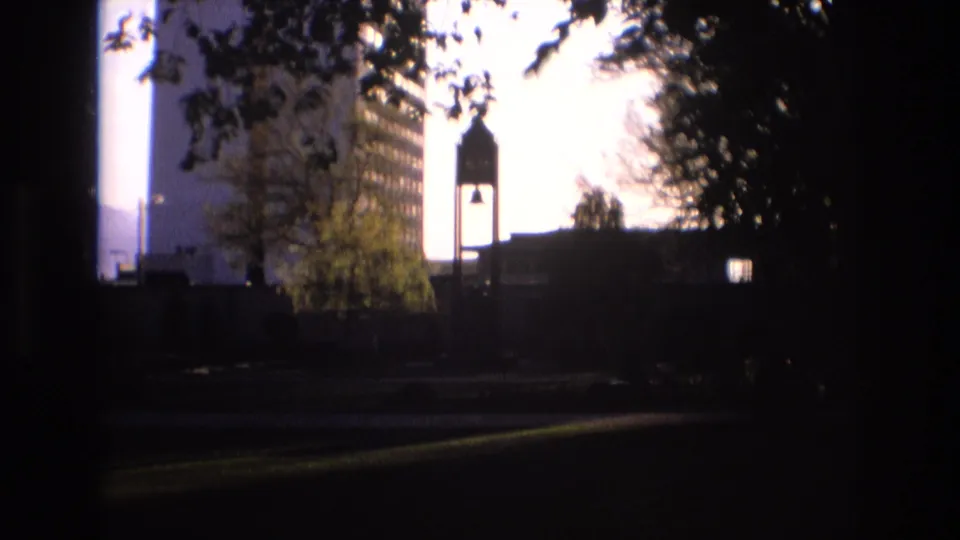 1973 Salt Lake City frame showing a shaded foreground, leafy trees, a silhouetted tower form, a nearby building, and bright afternoon sky.