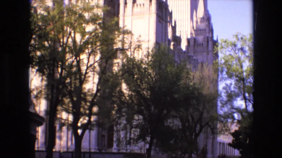 1973 Salt Lake City home-movie frame showing the Salt Lake Temple exterior partly obscured by trees under a clear bright sky.
