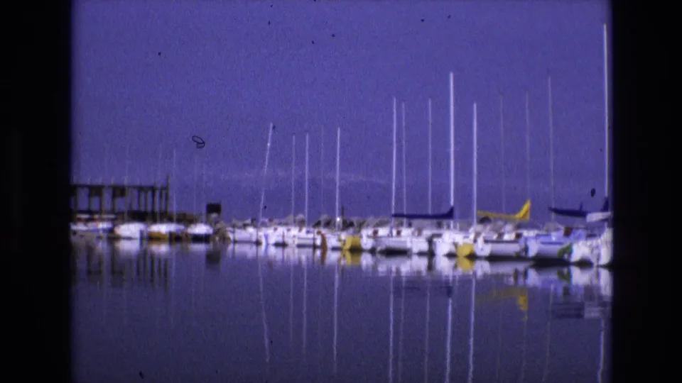 1973 Salt Lake City home-movie frame showing sailboats moored at a marina, their masts reflected across calm blue water.
