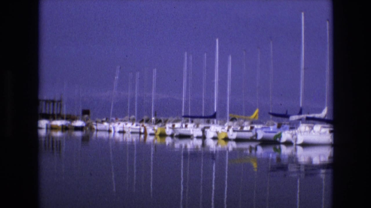 1973 Salt Lake City, Utah 8mm frame showing moored sailboats in still marina water, with tall mast reflections beneath a blue sky and low mountains in the distance.