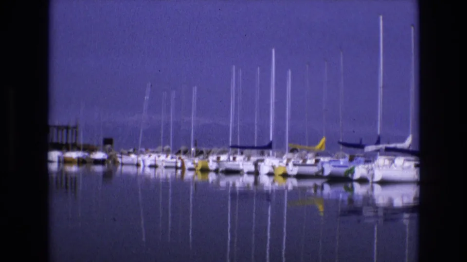 1973 Salt Lake City, Utah 8mm frame showing moored sailboats in still marina water, with tall mast reflections beneath a blue sky and low mountains in the distance.