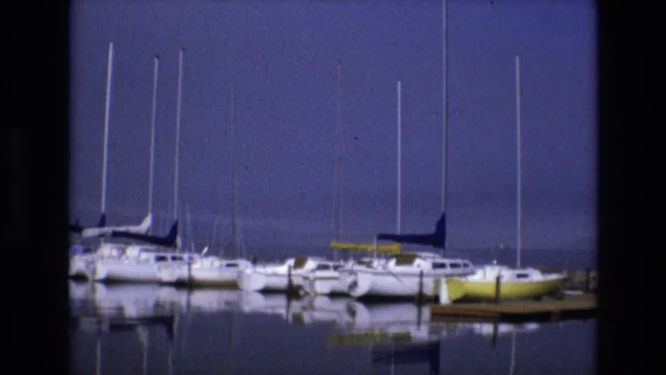 1973 8mm frame showing moored sailboats near dock posts, with a yellow boat at the right edge and mirrored masts in the calm marina water.