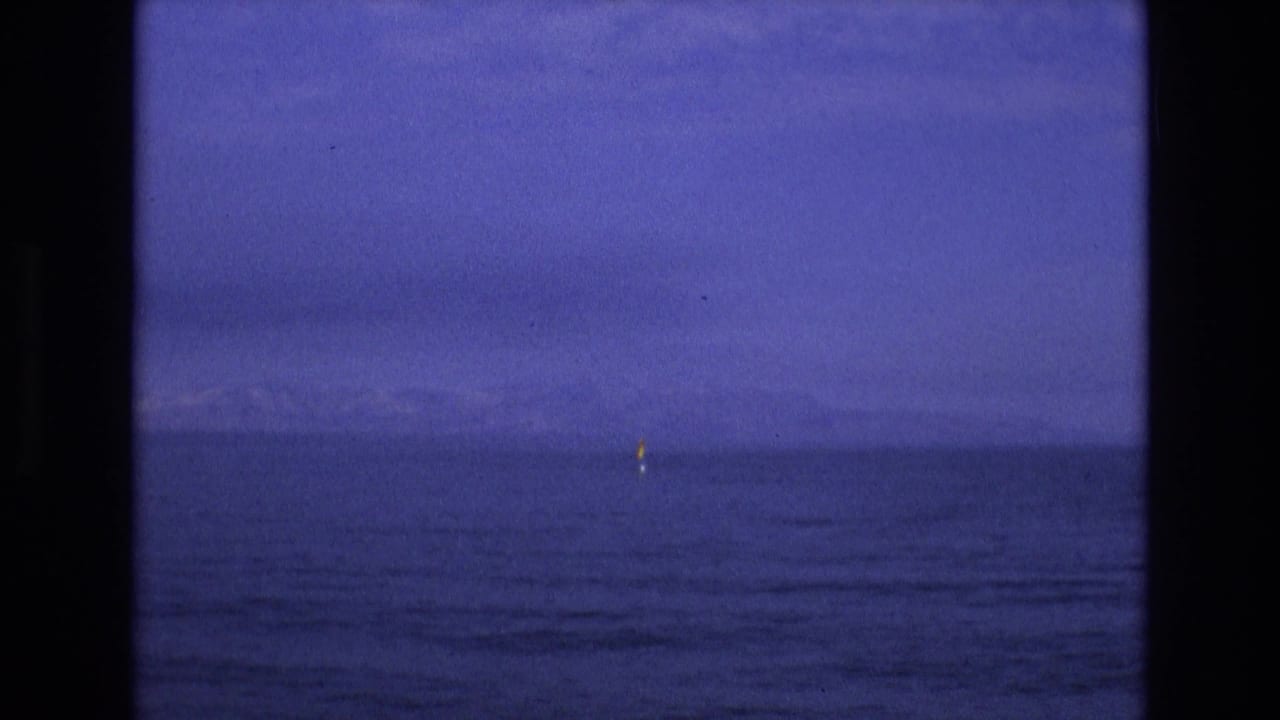 1973 8mm home-movie frame tied to Salt Lake City, Utah metadata, showing a tiny yellow sail or sailboat on calm water with faint mountains under a deep blue sky.