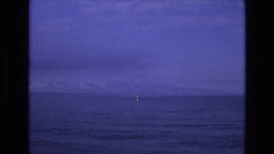 1973 8mm home-movie frame tied to Salt Lake City, Utah metadata, showing a tiny yellow sail or sailboat on calm water with faint mountains under a deep blue sky.