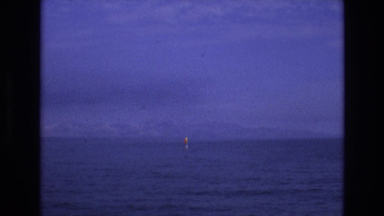 Small yellow sail near the horizon on dark blue water with faint mountains behind it.