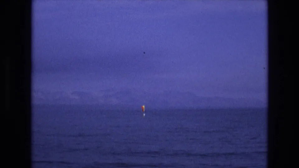 Soft 8mm frame from the supplied 1973 Salt Lake City, Utah clip showing a distant yellow sail on calm water, hazy mountains, and a pronounced blue-purple film cast.